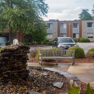 A small fountain and bench situated in front of an apartment complex parking lot.