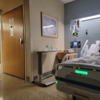 A view of a hospital patient room, showcasing the bedside area with medical equipment and access to the hallway.