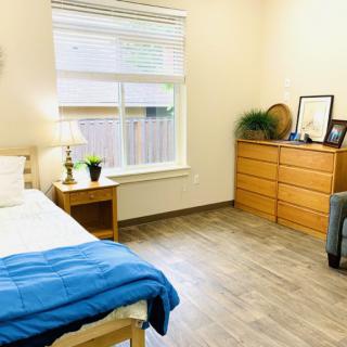 A minimalist bedroom featuring a cozy bed, wooden dresser, and natural light from a large window.