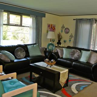 A cozy living room with dark sofas, colorful pillows, and a central coffee table, featuring decorative wall art and large windows.