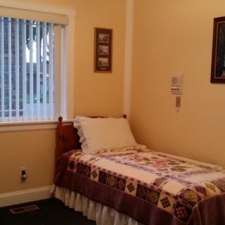A cozy bedroom with a single bed covered in a quilt, featuring framed pictures and a window with blinds.