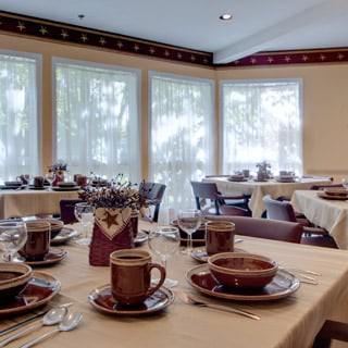 A cozy dining room with neatly set tables, featuring brown cups and plates near large windows.