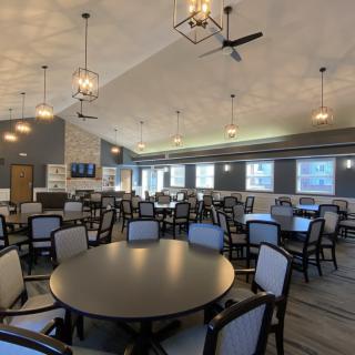A spacious dining area with multiple tables and chairs, elegantly lit by modern chandeliers, featuring large windows and ceiling fans.