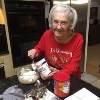 An elderly woman in a red sweatshirt is joyfully preparing food in the kitchen.