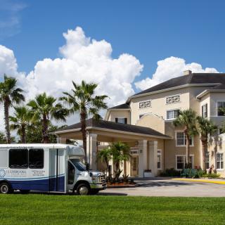 A building with a covered entrance surrounded by palm trees and a vehicle parked nearby under a blue sky with clouds.