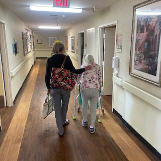 A caregiver gently assists an elderly woman as they walk down a corridor.