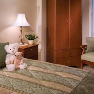 A cozy bedroom featuring teddy bears on a neatly made bed with green and beige bedding, a lamp on the nightstand, and a wardrobe in the background.