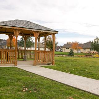 A wooden gazebo in a park with a grassy landscape and a concrete pathway.