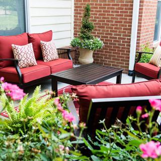 A cozy outdoor patio setup with red cushioned seating and a coffee table surrounded by lush greenery and flowers.
