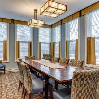 A well-lit dining room with a long table and patterned chairs, surrounded by large windows with curtains.