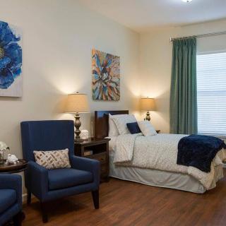 A cozy and elegantly decorated bedroom featuring a bed, two blue armchairs, artwork on the walls, and soft ambient lighting.