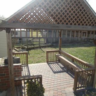 A wooden pergola with a lattice roof providing shade over a brick patio, featuring built-in benches and an open view to the backyard.