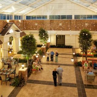 A spacious indoor atrium with people walking and conversing, surrounded by greenery and elegant architectural elements.