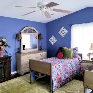 A cozy bedroom with blue walls, featuring a single bed adorned with floral bedding and decorative pillows. The room includes a dresser with a mirror, a ceiling fan, and various decorative elements.