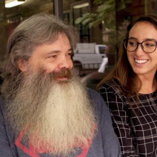 A man with a long beard and a woman wearing glasses share a joyful moment outdoors.