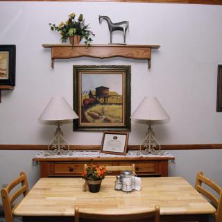 A cozy dining room setup featuring a wooden table and chairs, adorned with a central floral arrangement and salt and pepper shakers. The background showcases a painting flanked by two lamps, with a shelf above displaying decorative items including a horse