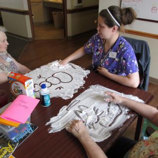 A caregiver engages seniors in an art therapy session using shaving cream as a creative medium.