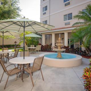 A cozy outdoor patio area with tables, chairs, and umbrellas surrounding a central fountain, adjacent to a building with palm trees providing shade.