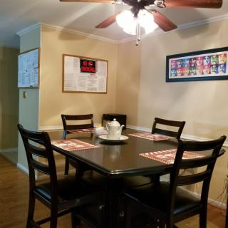 A cozy dining room featuring a dark wooden table with four chairs, adorned with placemats and a centerpiece, under a ceiling fan and surrounded by artwork on the walls.