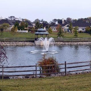 A serene park scene with a fountain in a lake, surrounded by a wooden fence and trees, with houses in the background.