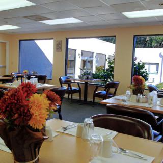A bright dining area with tables set for a meal, featuring floral decorations and large windows.