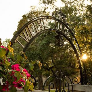 A decorative archway reading 'Twelve Oaks' adorned with flowers, set against a backdrop of trees and a sunset.