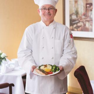A chef in a white uniform presenting a plated dish at a restaurant.