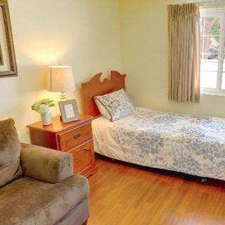 A cozy bedroom with a single bed, a nightstand with a lamp, and a comfortable chair by the window.