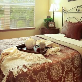 A cozy bedroom with a neatly made bed, adorned with a patterned blanket and pillows, featuring a tray with cups and a teapot. Soft lighting from a lamp beside the window complements the warm decor.