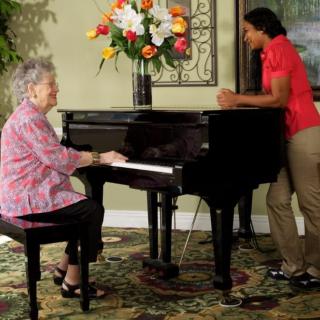 An elderly woman playing the piano while engaging in a conversation with another person standing nearby.