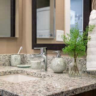 A tidy bathroom countertop featuring a granite surface, decorative jars, fresh greenery, and neatly stacked towels beside a mirror.