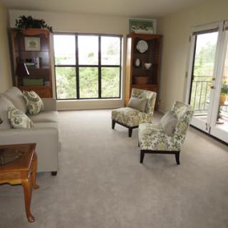 A cozy living room featuring a sofa, two chairs, and bookshelves, with a large window and sliding door leading to a balcony.