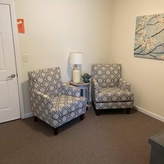 A cozy living room corner featuring two patterned chairs, a small side table with a lamp, and a floral artwork on the wall.