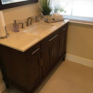 A bathroom vanity with a sink, faucet, and cabinet under a mirror, featuring a plant and tissues by the window.