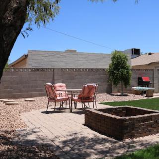 A cozy backyard patio with chairs and a table situated near a fire pit, surrounded by trees and a fence.