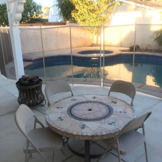 A backyard patio with a round table, chairs, and a view of the swimming pool enclosed by a safety fence.