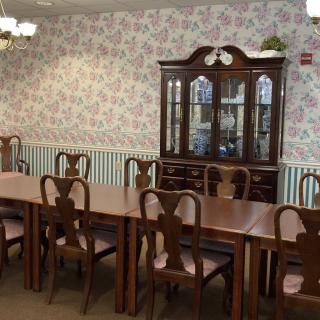 A classical dining room with a long wooden table, chairs, and a display cabinet against floral wallpaper.