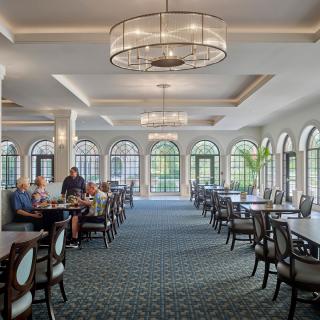 A spacious dining area with elegant chandeliers and large arched windows letting in natural light, featuring neatly arranged tables and chairs.