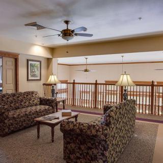 A cozy living room area with patterned sofas, a coffee table, and warm lighting from standing lamps. A ceiling fan is installed above, and there's a wooden railing providing a balcony view.