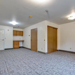 A spacious empty room with a kitchenette and carpeted floor.