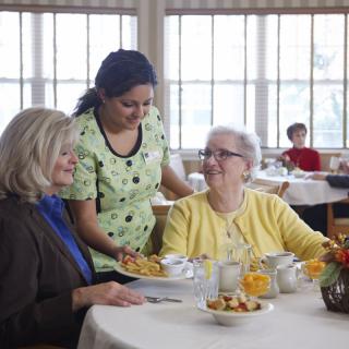 A caregiver interacting with two elderly women in a nursing home dining room.
