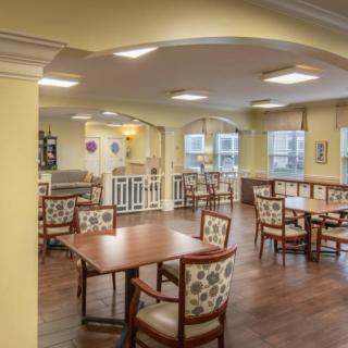 A well-lit dining area with multiple tables and chairs, featuring large windows and wooden flooring.
