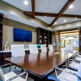 A modern conference room with a large meeting table, white chairs, and a wall-mounted TV screen.