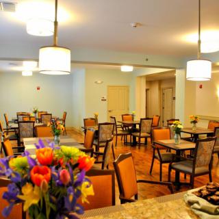 A brightly lit dining area with multiple tables and chairs, adorned with colorful flowers.