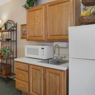 A small kitchen area with wooden cabinets, a white microwave and refrigerator, and a metal shelf.