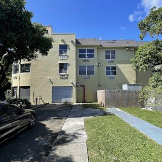 A two-story yellow apartment building with a parking area and landscaped lawn under a clear blue sky.