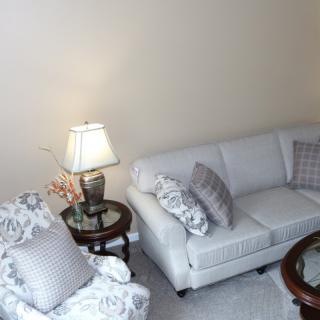 A cozy living room with a white sofa and matching armchair, adorned with patterned cushions and accompanied by a side table with a lamp.