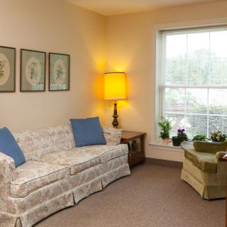 A cozy living room with a floral sofa, armchair, and warm lighting from a lamp beside the window.