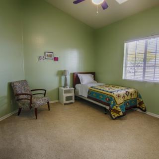 A minimalist bedroom with green walls and a single bed, featuring a chair and a lamp by the window.