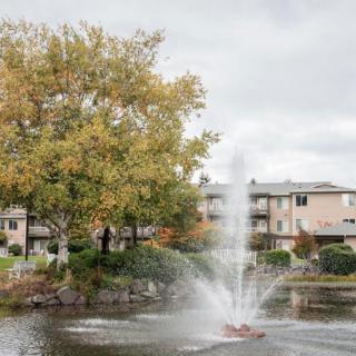 A tranquil pond with a fountain, surrounded by apartment buildings and autumn foliage under a cloudy sky.